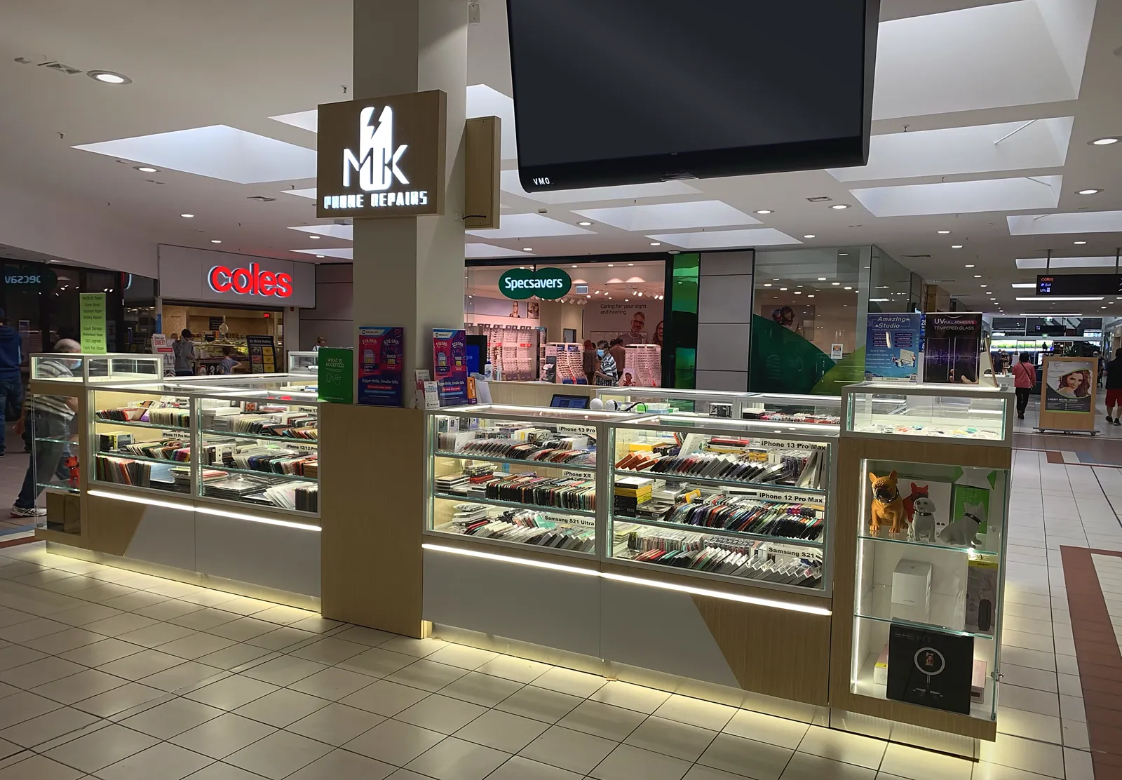 MK Phone Repairs kiosk at Brandon Park Shopping Centre, with glass cabinets of phone cases and the lit MK sign, near Coles and Specsavers